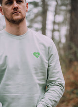Man wearing a Tree Love EMB Sweatshirt - an organic cotton sweatshirt in pale green with an embroidered tree and heart graphic from Sutsu.