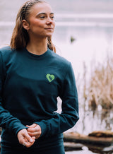 Woman wearing a Tree Love EMB Sweatshirt - an organic cotton sweatshirt in teal with an embroidered tree and heart graphic from Sutsu.