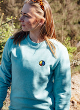 Woman wearing a light blue Sutsu Simon Says Peace EMB Sweatshirt - organic cotton sweatshirts with embroidered multi-coloured peace symbol.